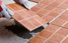 Persona con guantes de trabajo instalando una baldosa marrón sobre una superficie, junto a otras zonas alicatadas.