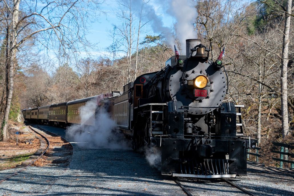 Steam locomotive pulling a vintage passenger train through a wooded autumn landscape