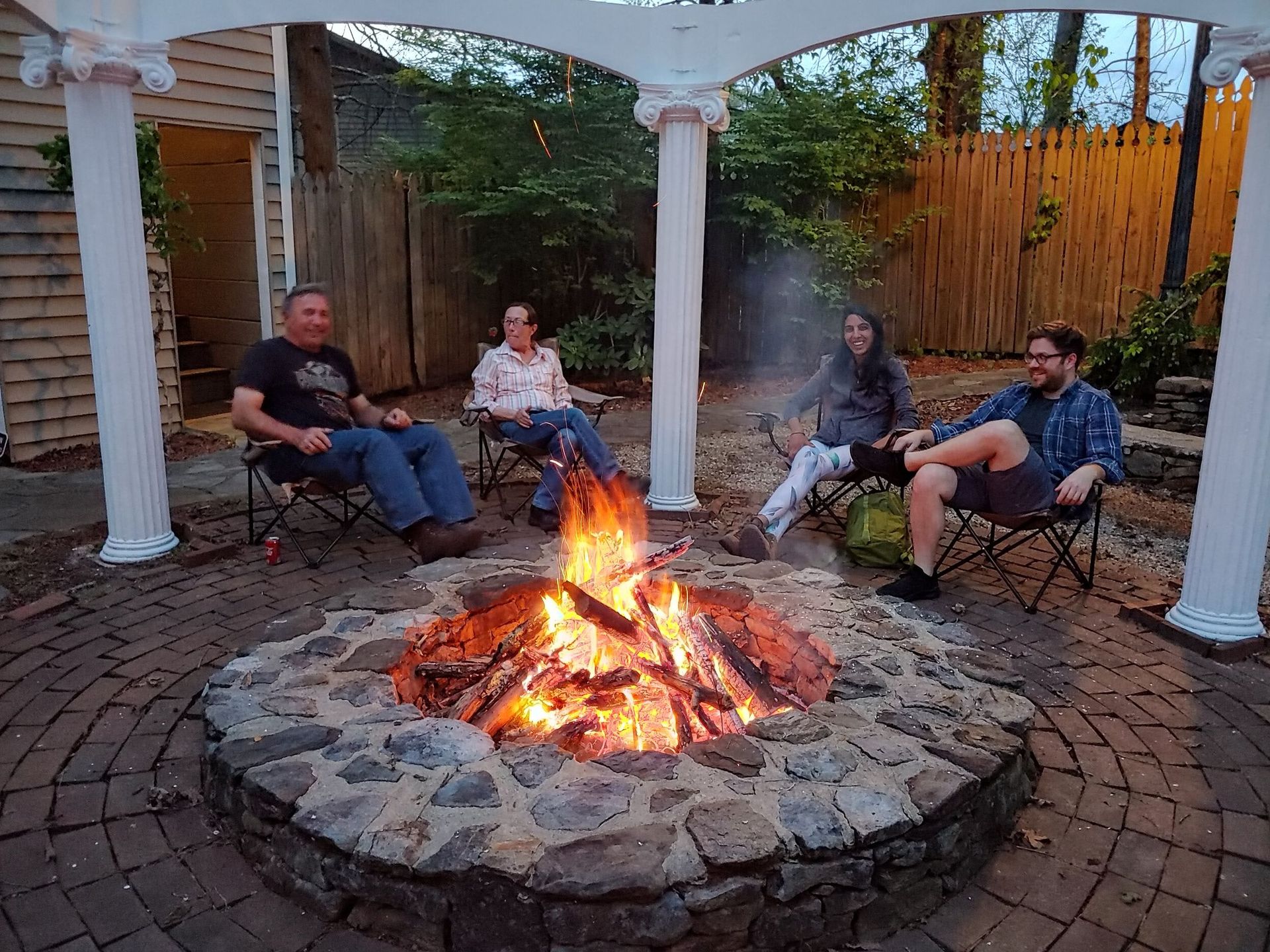 Four people sit around a stone fire pit under a white pergola in a backyard at dusk.