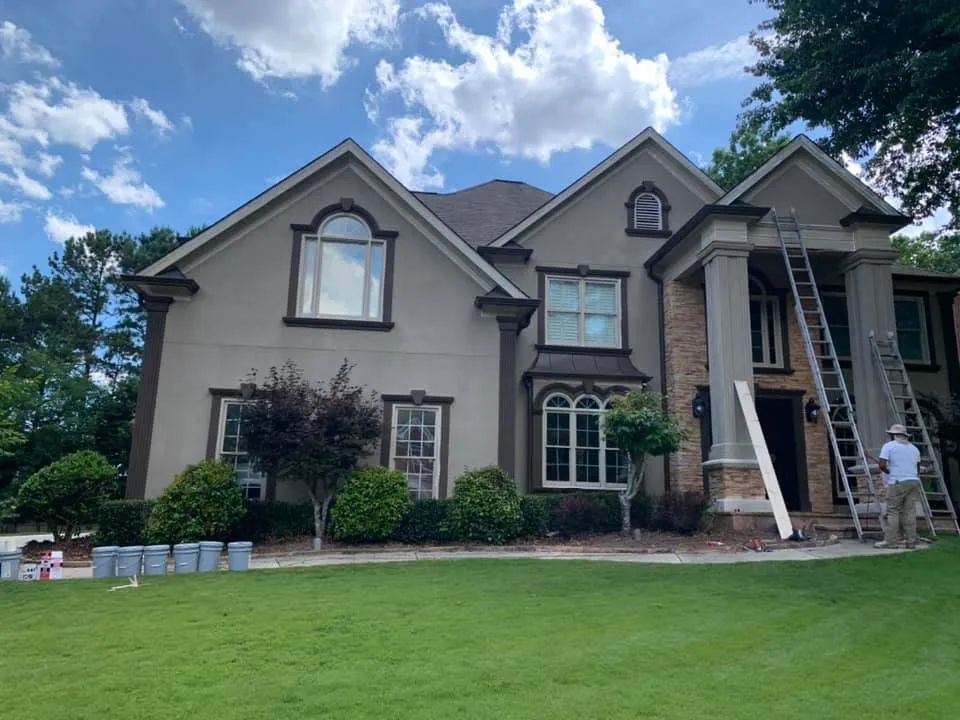 House exterior being painted; ladders, person, light-colored stucco, dark trim, blue sky.