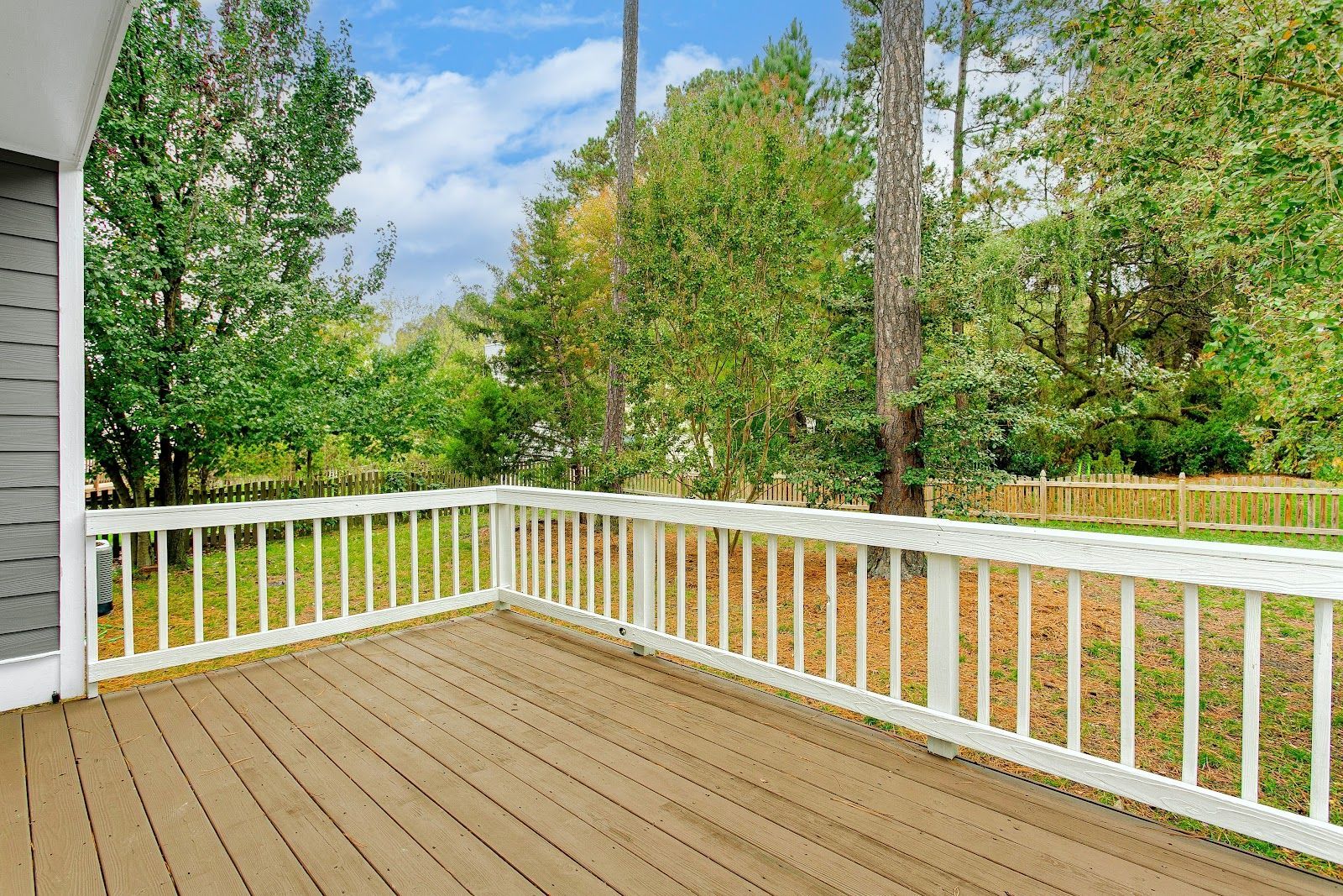 Wooden deck with white railing, surrounded by trees and greenery under a partly cloudy sky.