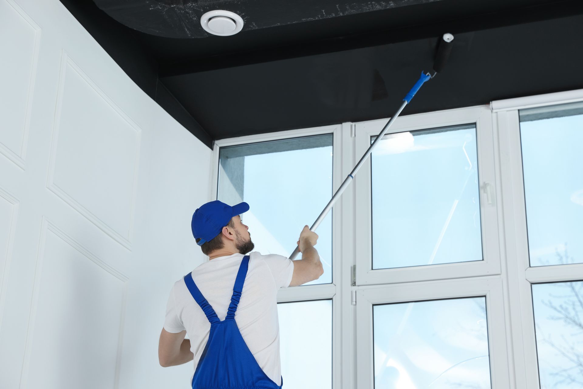 Person in blue overalls paints black ceiling with a roller, near a window.