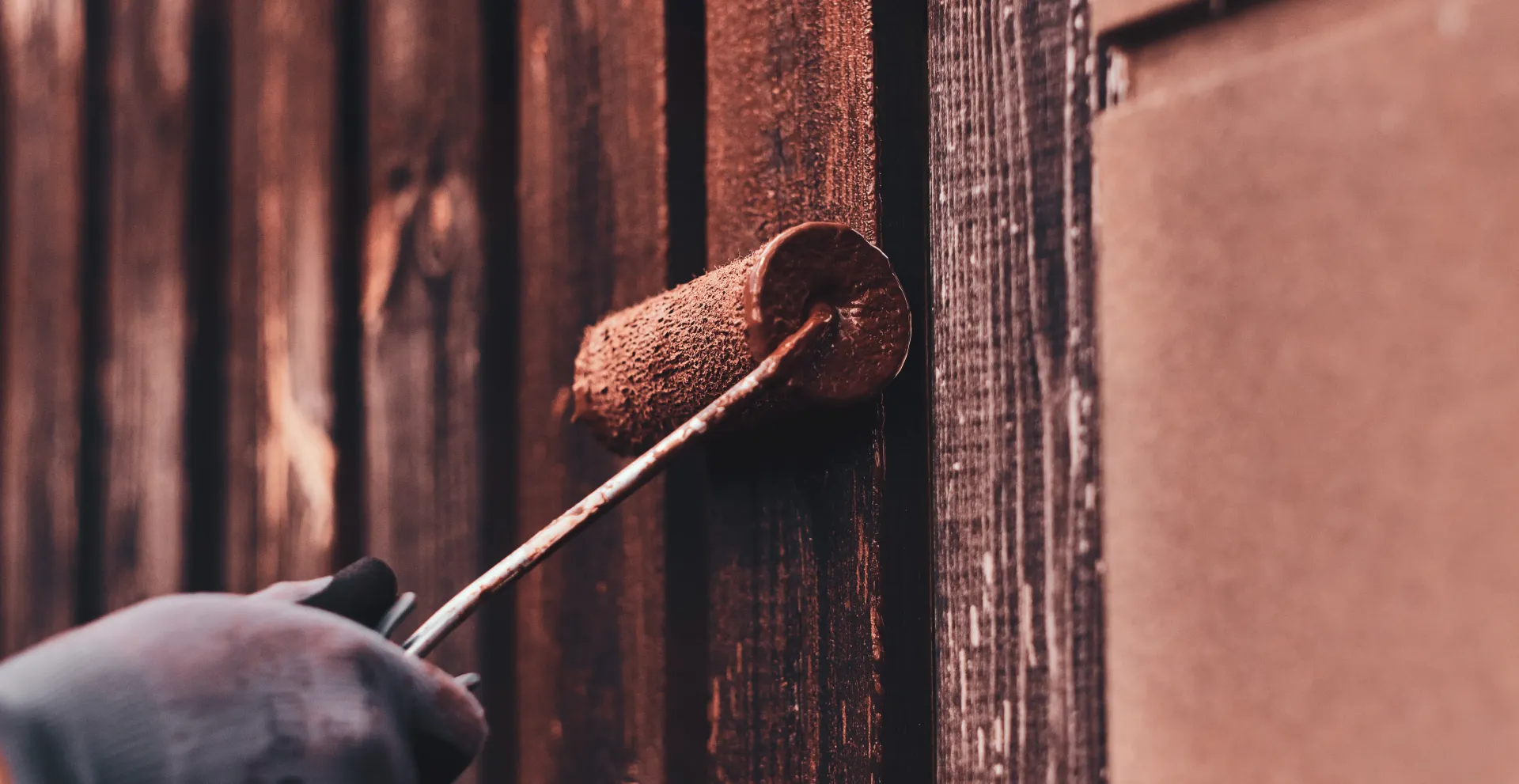 A gloved hand uses a paint roller to apply brown stain to wooden planks.