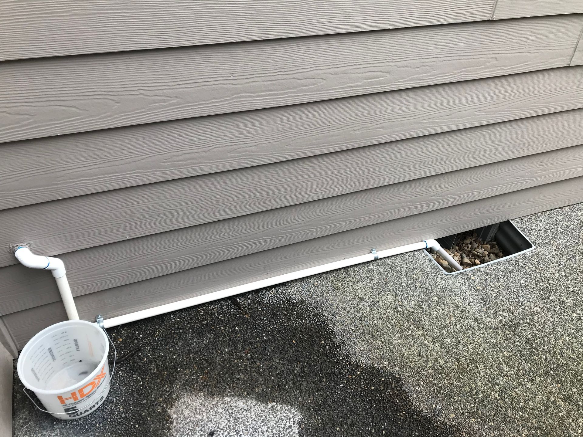 A white bucket is sitting next to a pipe on the side of a house.