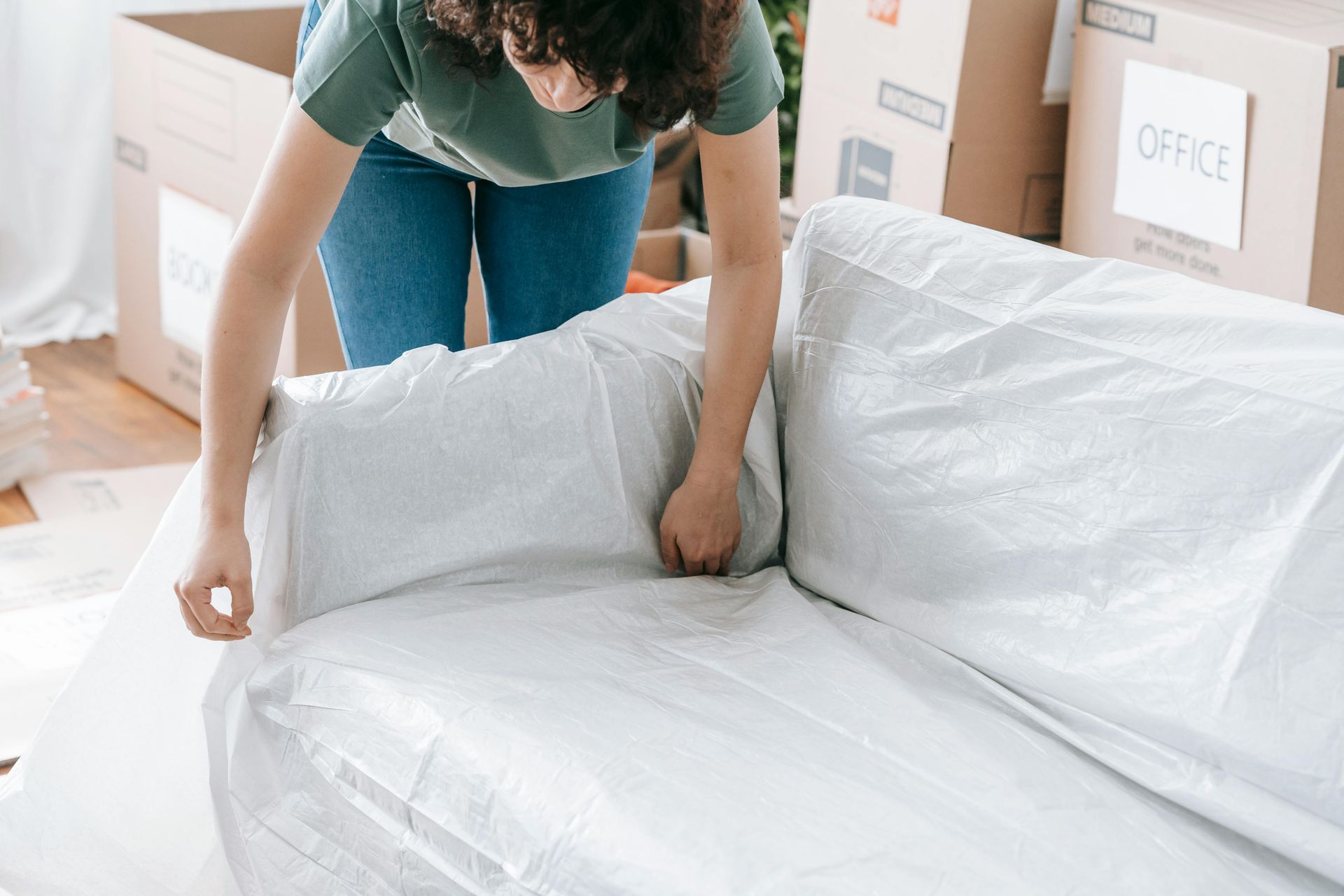 Woman wrapping a white sofa in plastic during a move, surrounded by moving boxes.