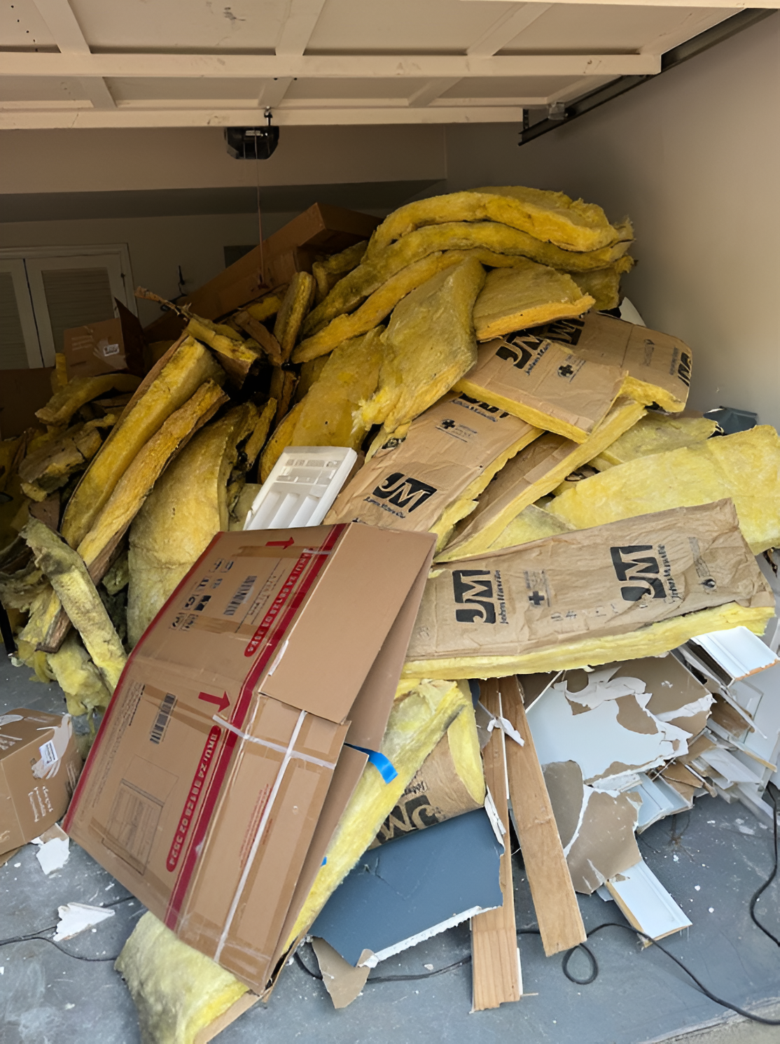 Pile of construction debris in a garage, including yellow insulation, cardboard boxes, and wood scraps.