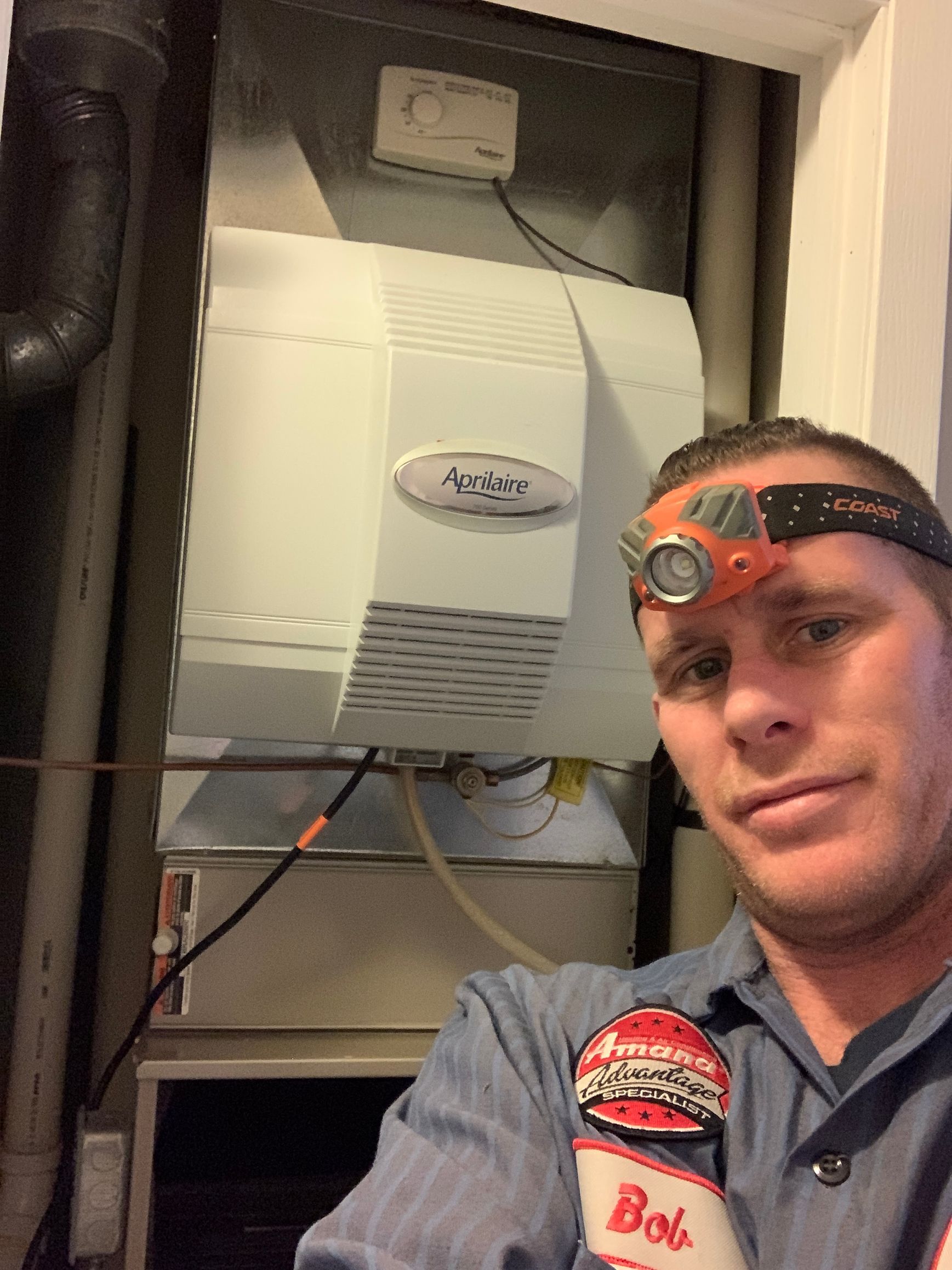 Man wearing a headlamp and work shirt beside a wall-mounted boiler in a utility closet
