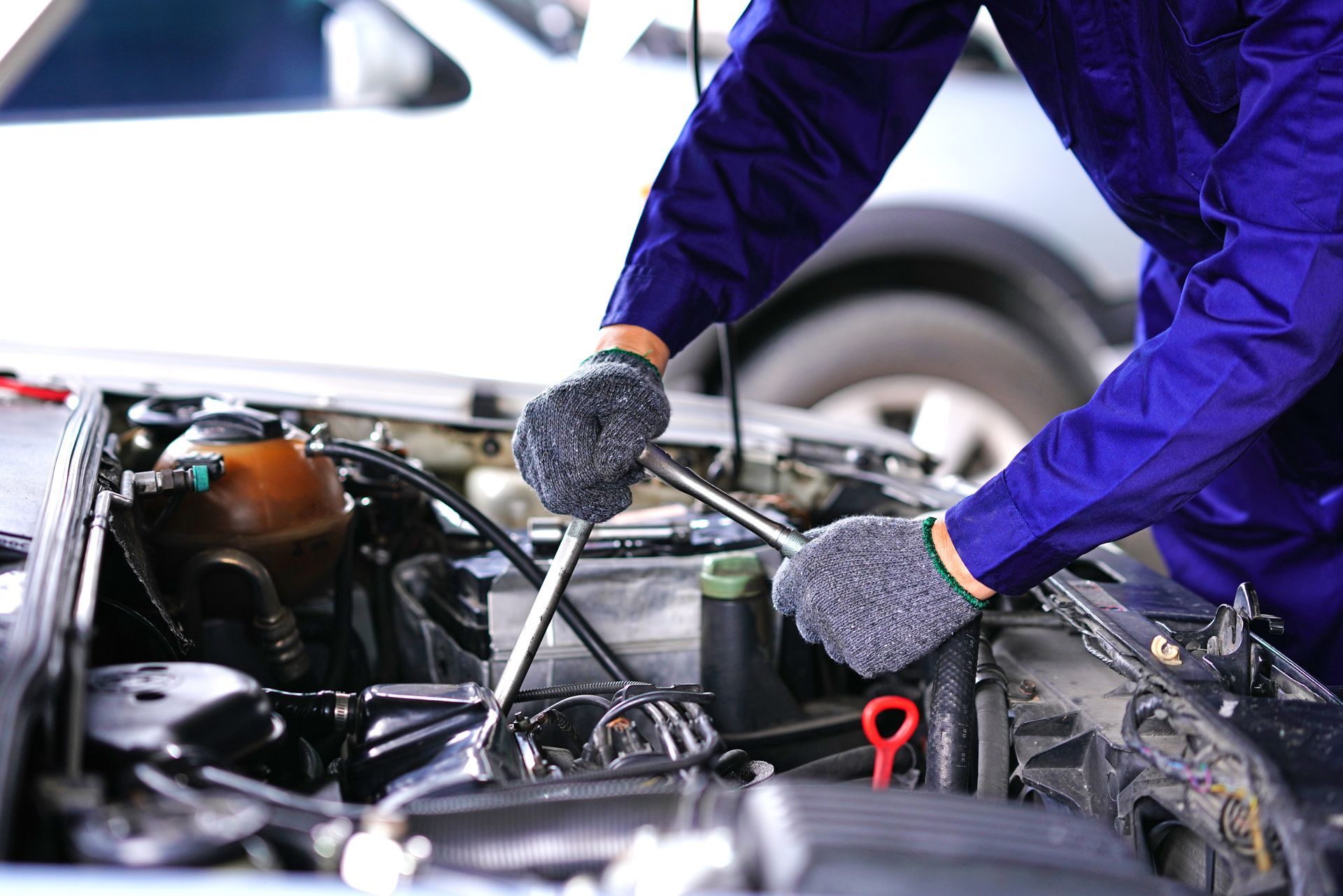Mechanic in blue overalls, working on a car engine with a wrench; gloves on.