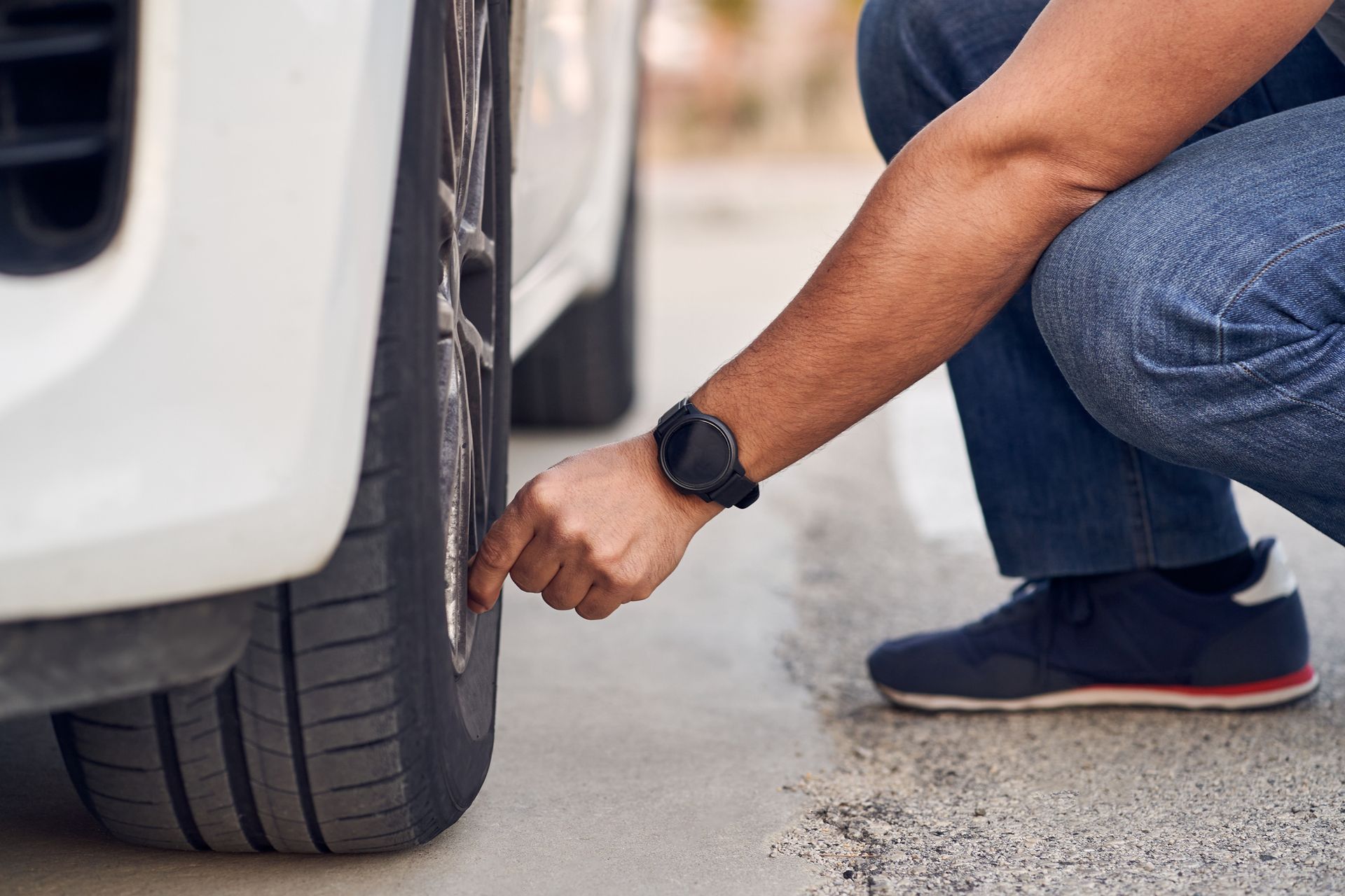 Person checks tire pressure of a white car, wearing blue jeans, watch, and sneakers outdoors.