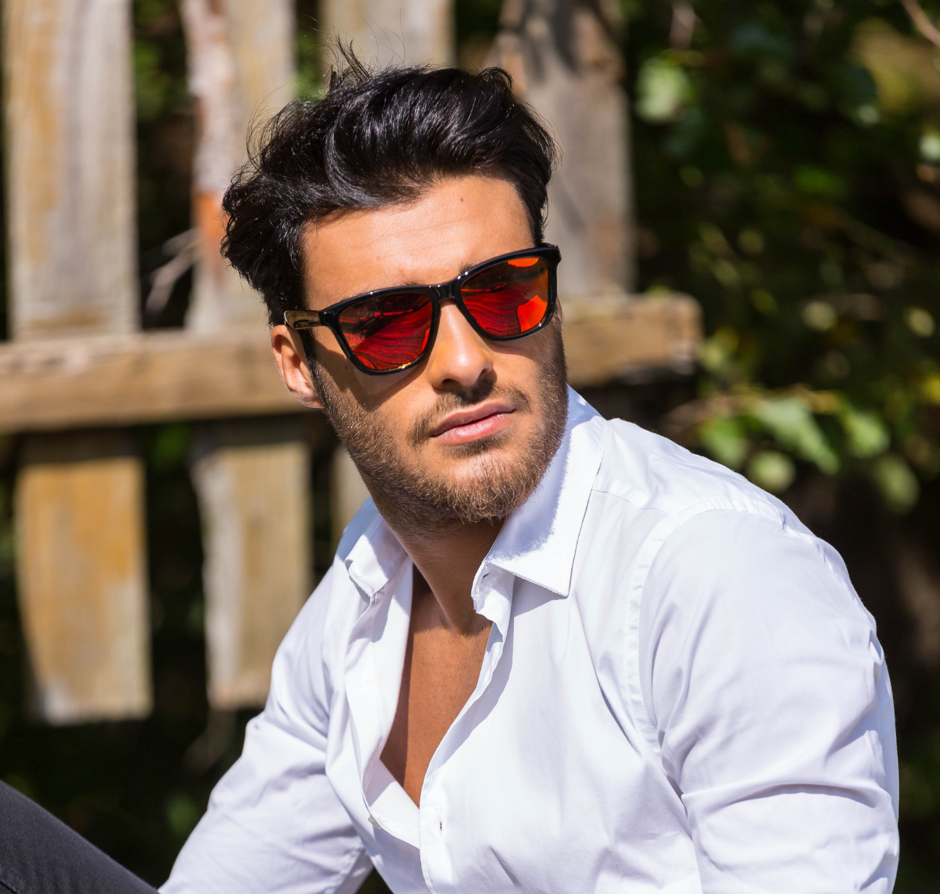 Un hombre que lleva gafas de sol y una camisa blanca está sentado frente a una valla de madera.