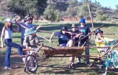 Group of kids posing with a wooden cart made of sticks, wheels, and bike parts in a sunny outdoor setting.