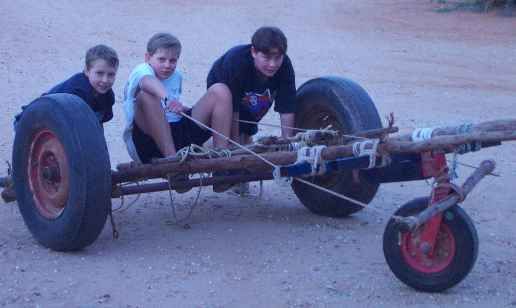 Three boys on a homemade wooden cart with large tires, outdoors on dirt, smiling.