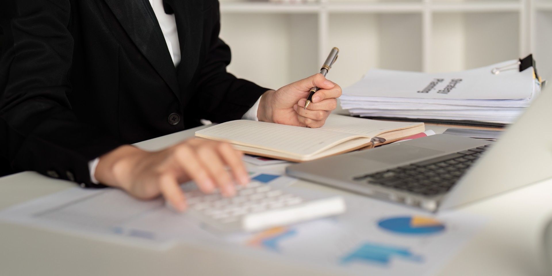 A woman is sitting at a desk using a calculator and writing in a notebook.
