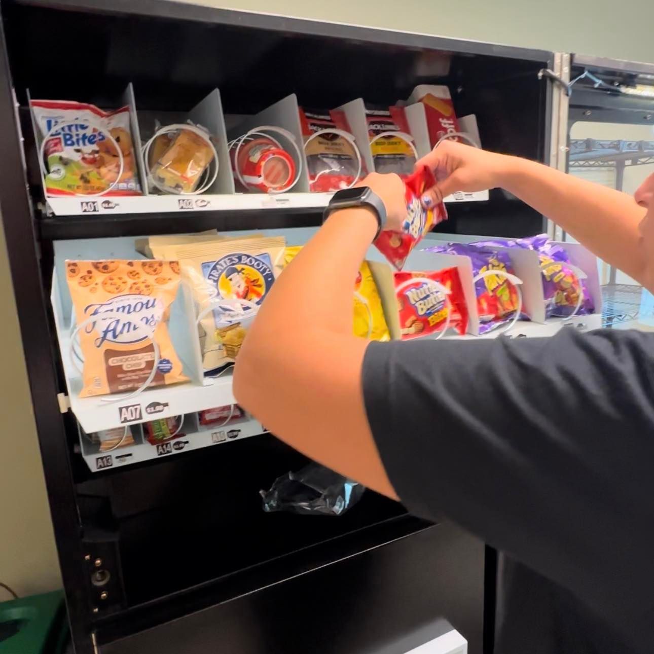A person’s arms are visible as they restock snacks into a black vending machine filled with assorted bagged goods.