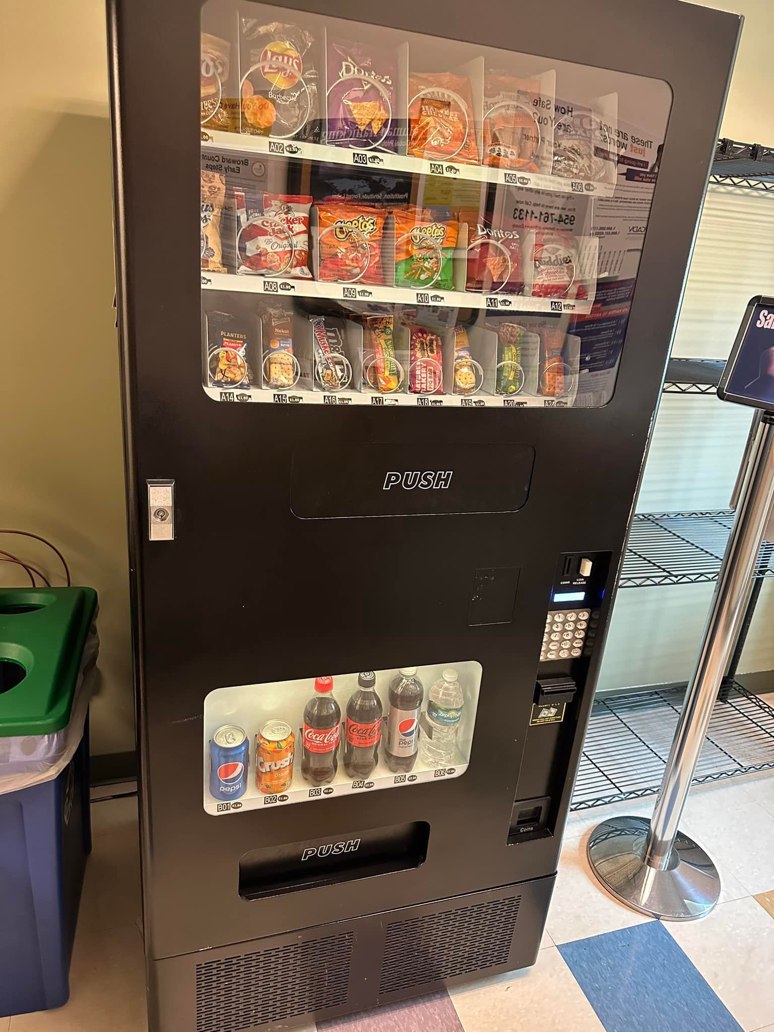 A black, floor-standing vending machine stocked with various snack bags on the top shelves and cold drinks on the bottom.