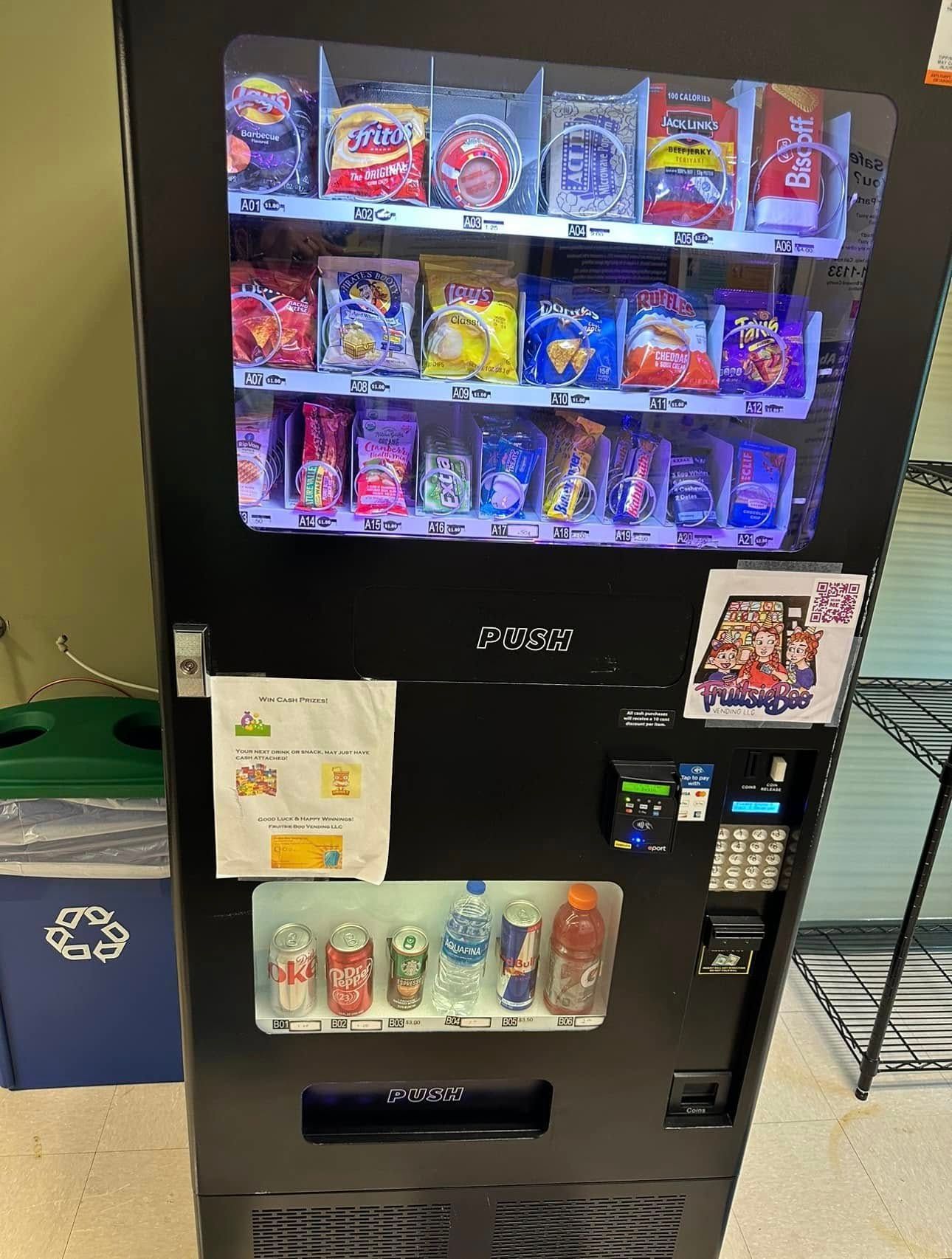 A black vending machine stocked with various snacks and drinks, standing in a room next to a blue recycling bin.