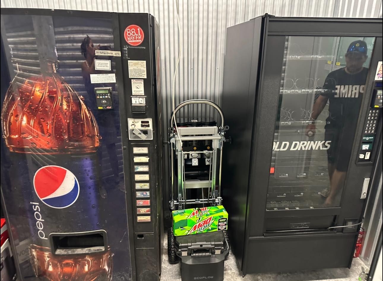 Two drink vending machines flanking a hand truck holding a case of Mountain Dew in a storage unit.