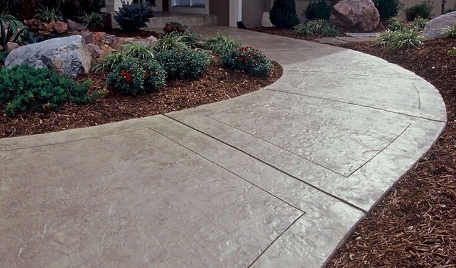A concrete walkway leading to a house surrounded by rocks and plants.