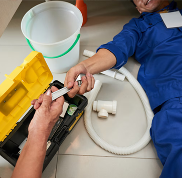 Plumber on floor, receiving tools from another hand, near toolbox and plumbing parts.
