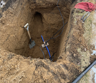 Blue pipe with valve in a dug-out hole with soil. Electrical wiring and metal components are visible.