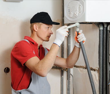 Plumber in red shirt, black cap, and gloves, working on a pipe in a construction setting.