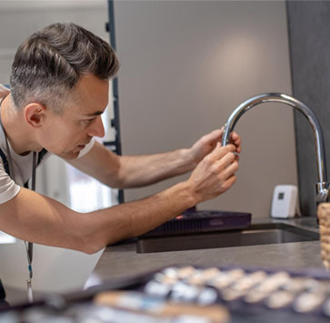 Man inspecting a kitchen faucet. He's leaning over the sink, focused, in a modern kitchen.