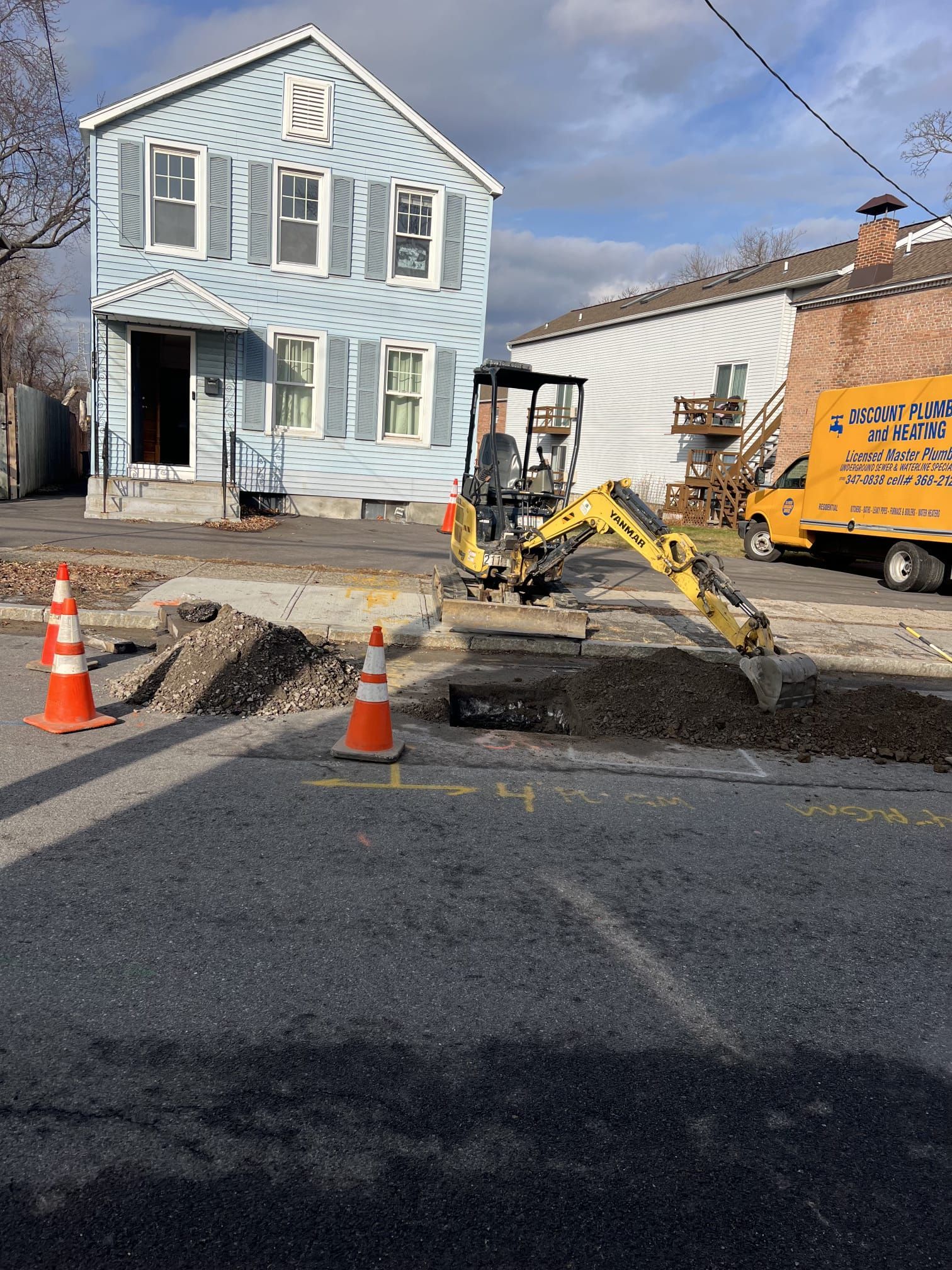 Mini excavator digs in a street next to a light blue house. Orange cones mark the work area. A yellow van is parked.