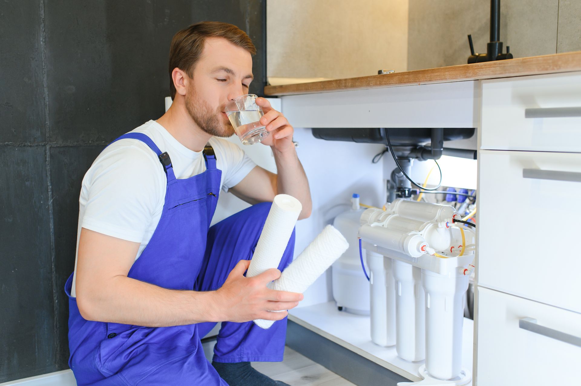 A man is drinking water from a glass while kneeling under a sink.