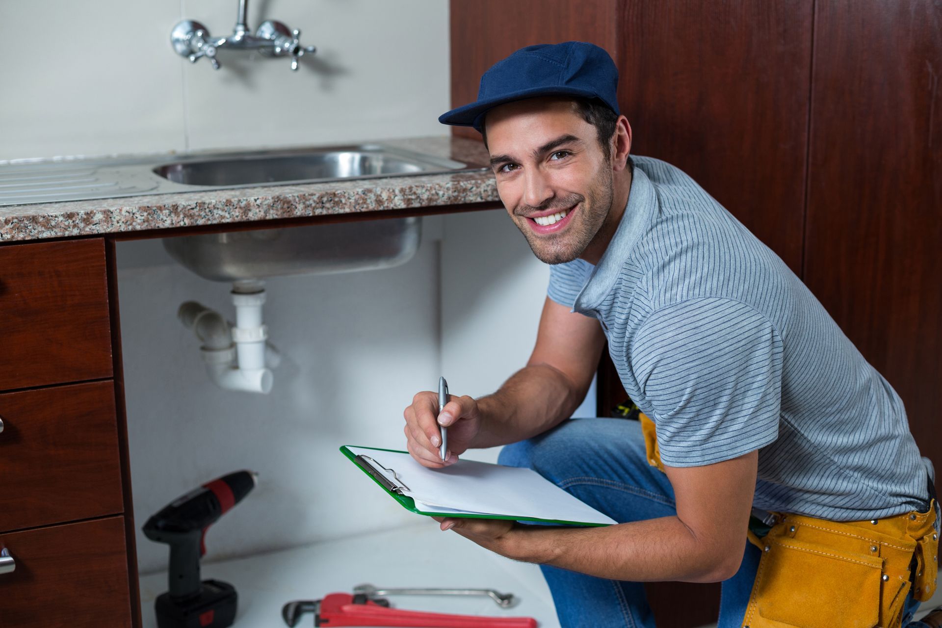A plumber is writing on a clipboard under a sink in a kitchen.