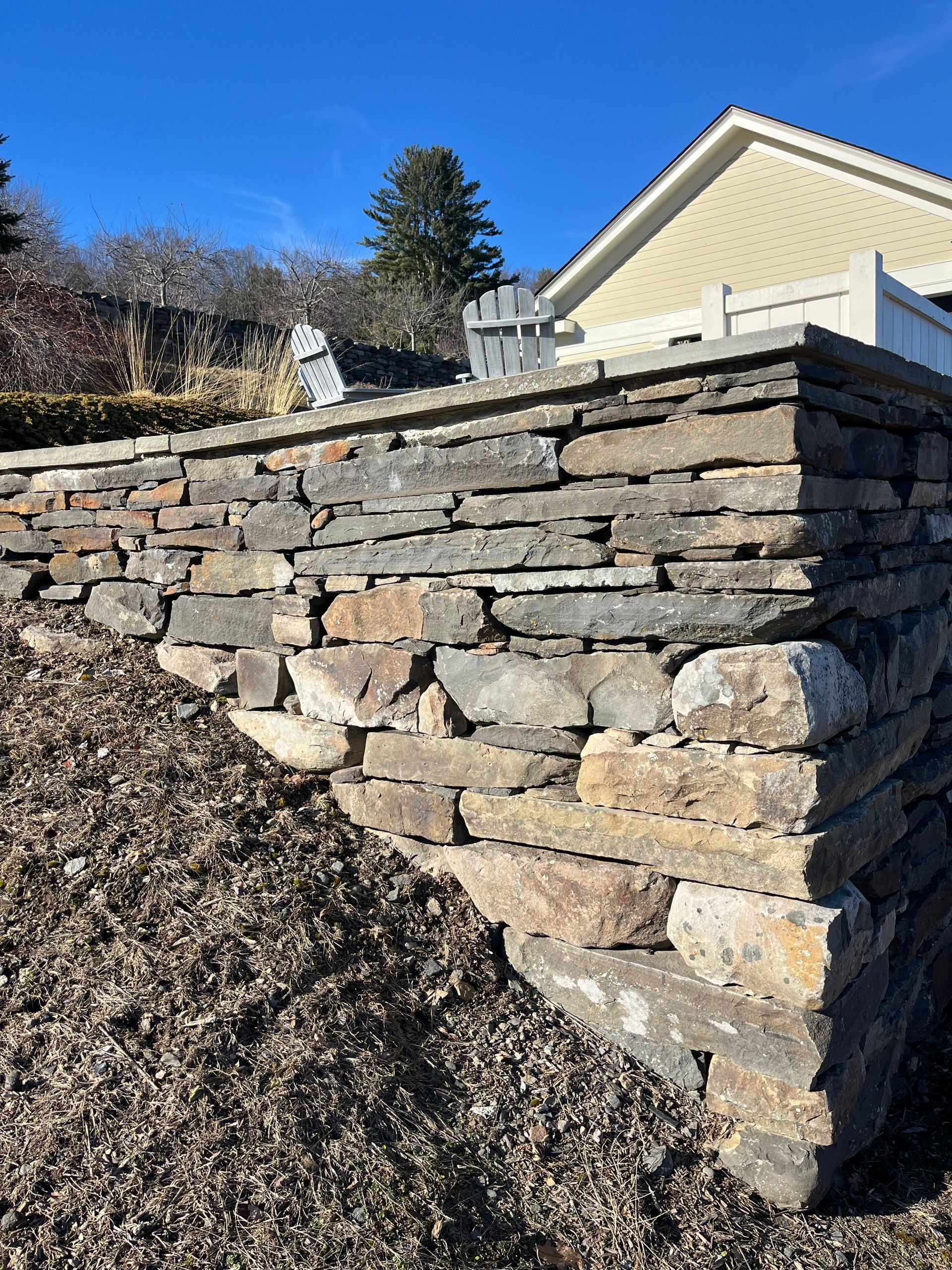A stone retaining wall built with horizontal slate pieces sits beside a sloped patch of mulch under a clear blue sky.