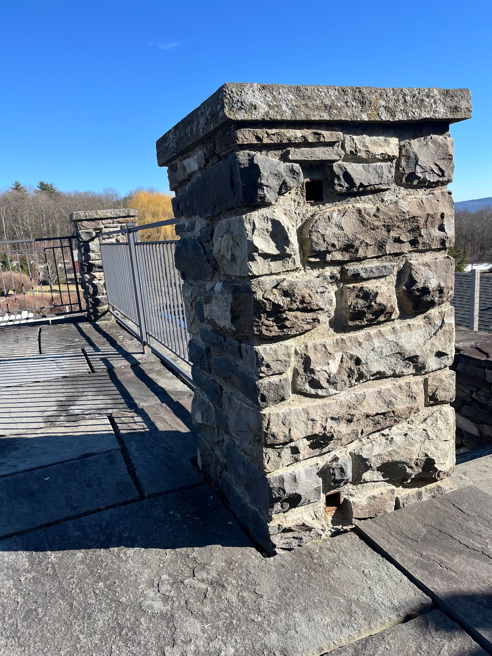 A stone pillar stands on a gray stone terrace under a clear blue sky, next to a metal fence railing.
