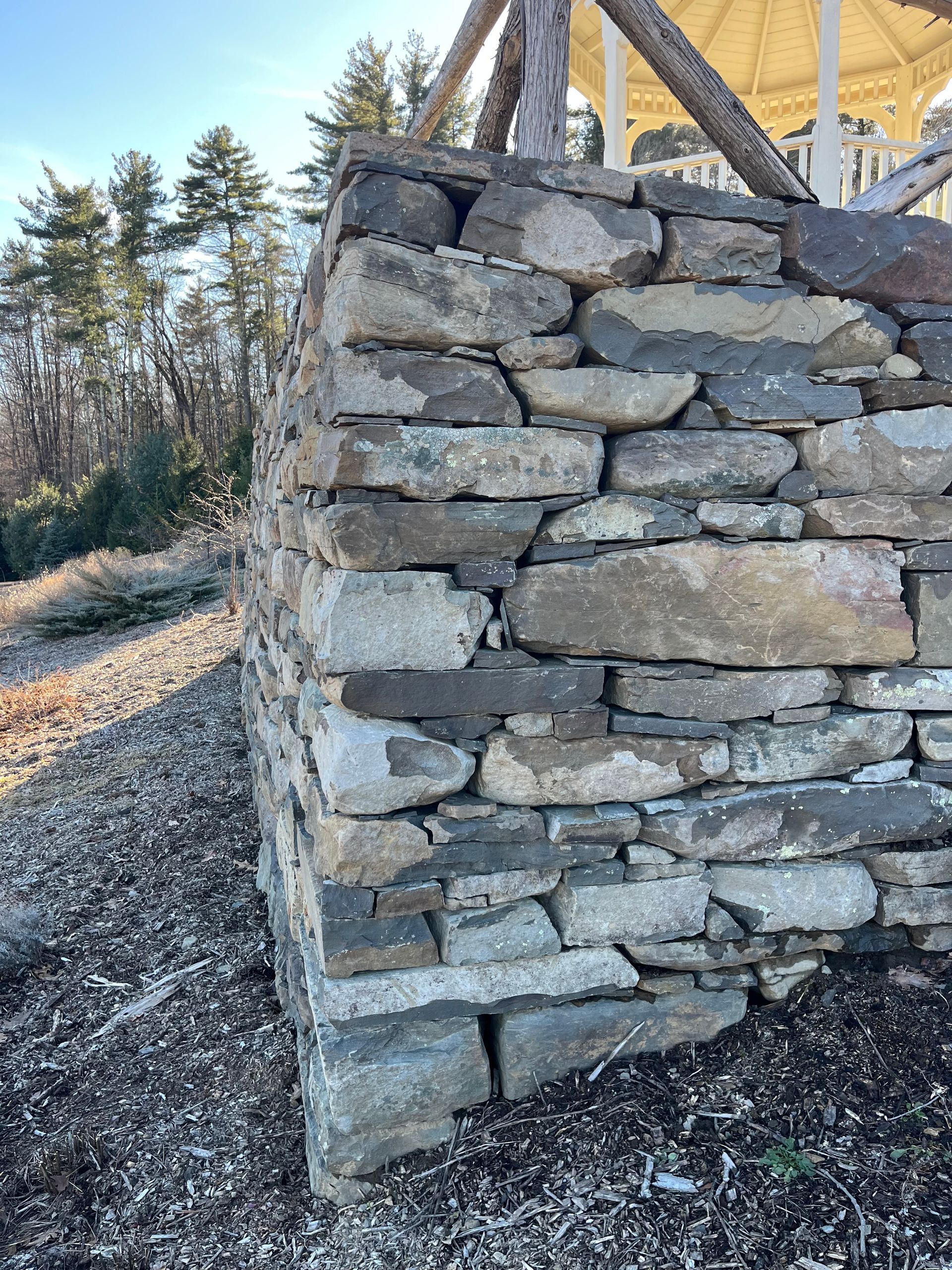A close-up view of a rustic, dry-stacked stone wall supporting a gazebo structure on a hill with pine trees in the background.