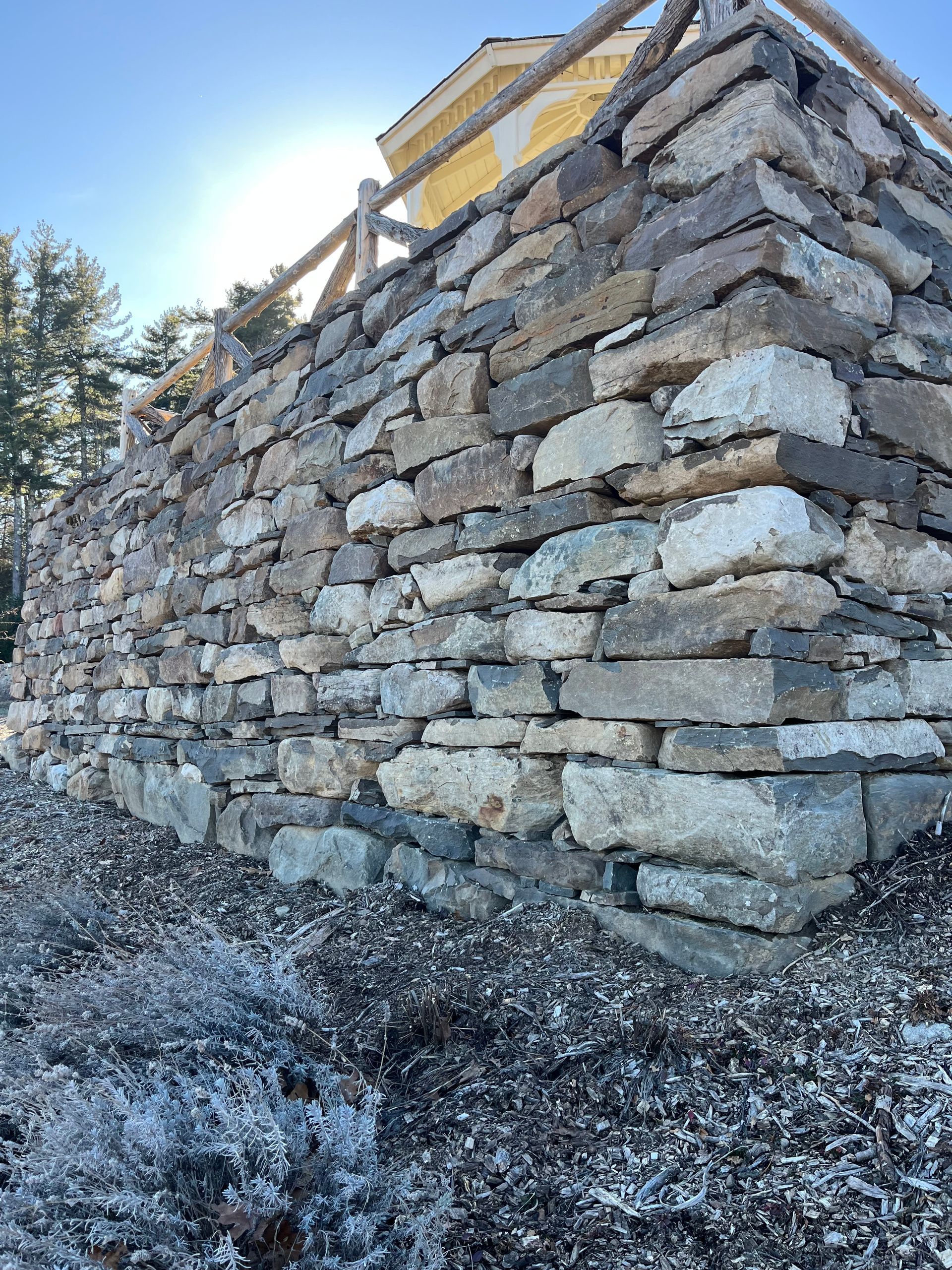 A low-angle view of a rustic, dry-stacked stone retaining wall with a wooden fence railing against a bright, sunny sky.