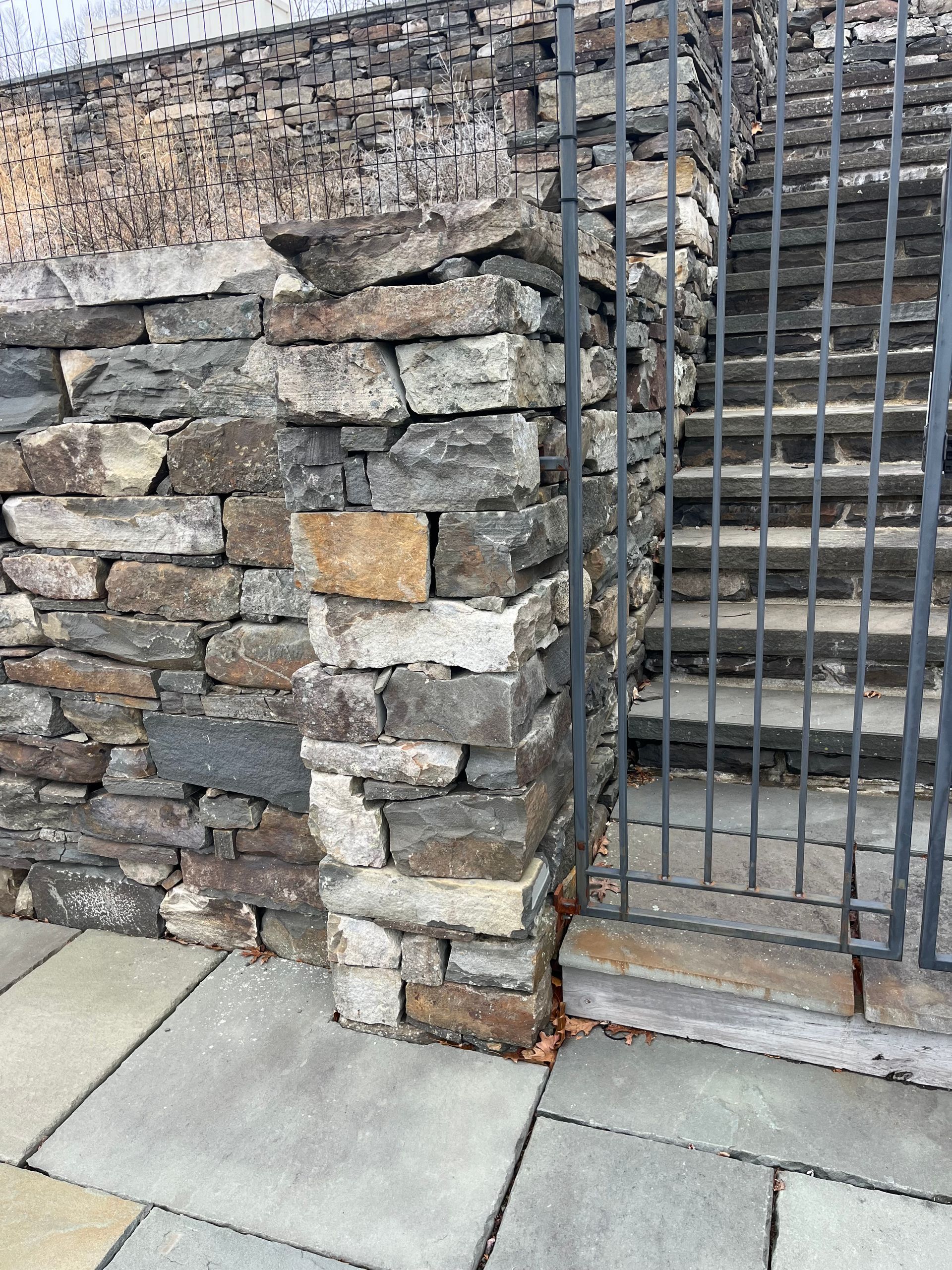 A stacked stone wall and pillar next to a gray metal gate and stone stairs.