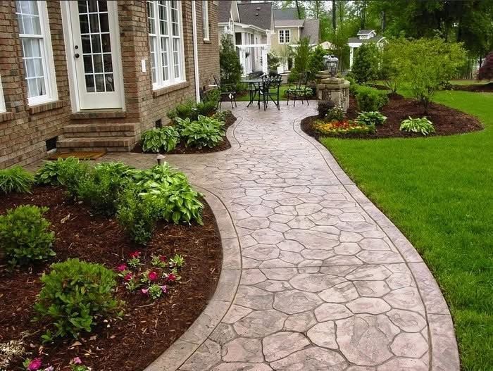 A stamped concrete walkway with a stone pattern curves past a brick house, mulched garden beds, and a lush green lawn.