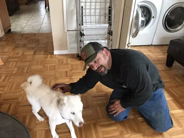 A man is kneeling down next to a white dog in a laundry room in South Bay, CA