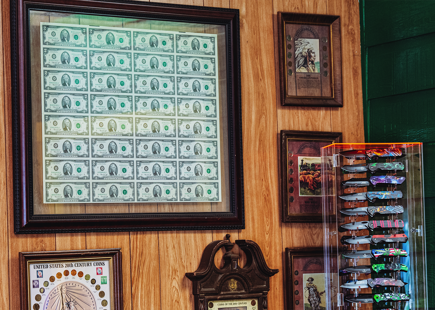 Wall display with framed dollar bills, artwork, clock, and knives in a lighted cabinet.
