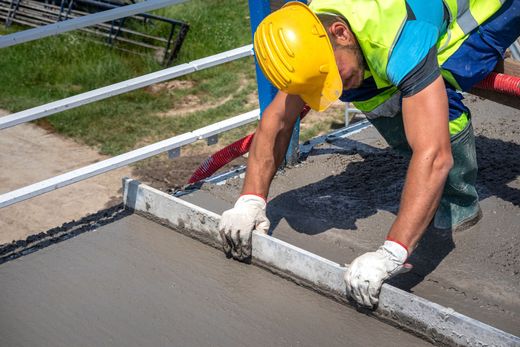 A concrete contractor with a yellow helmet is straightening and smoothing fresh driveway.