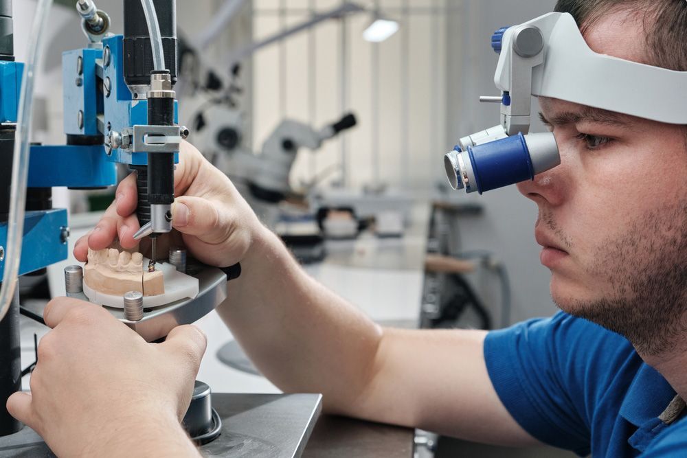 A Man In A Blue Shirt Is Working On A Denture Model Using A Machine — Dentures in Long Jetty, NSW