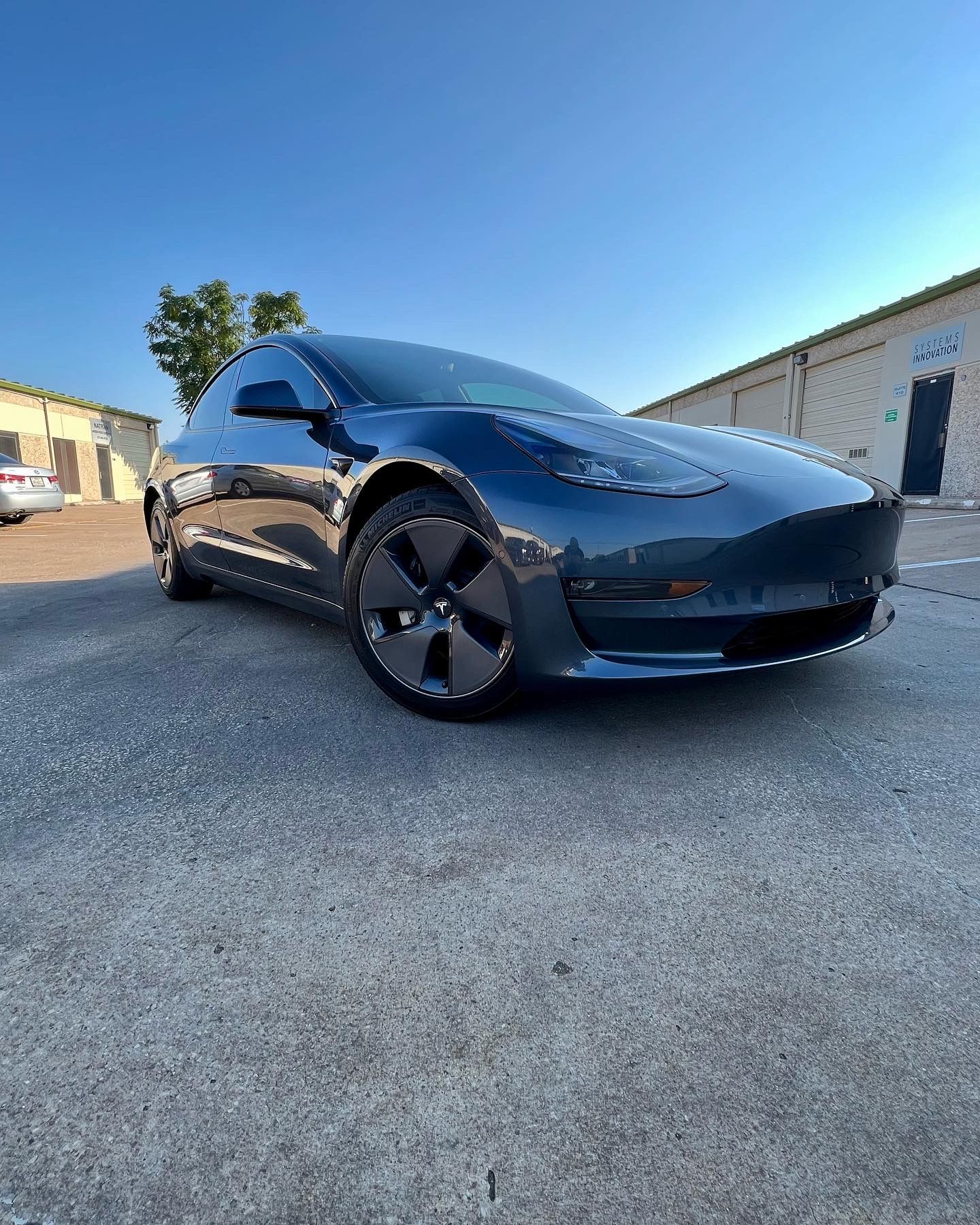 Blue Tesla Model 3 car parked on pavement under a clear blue sky.