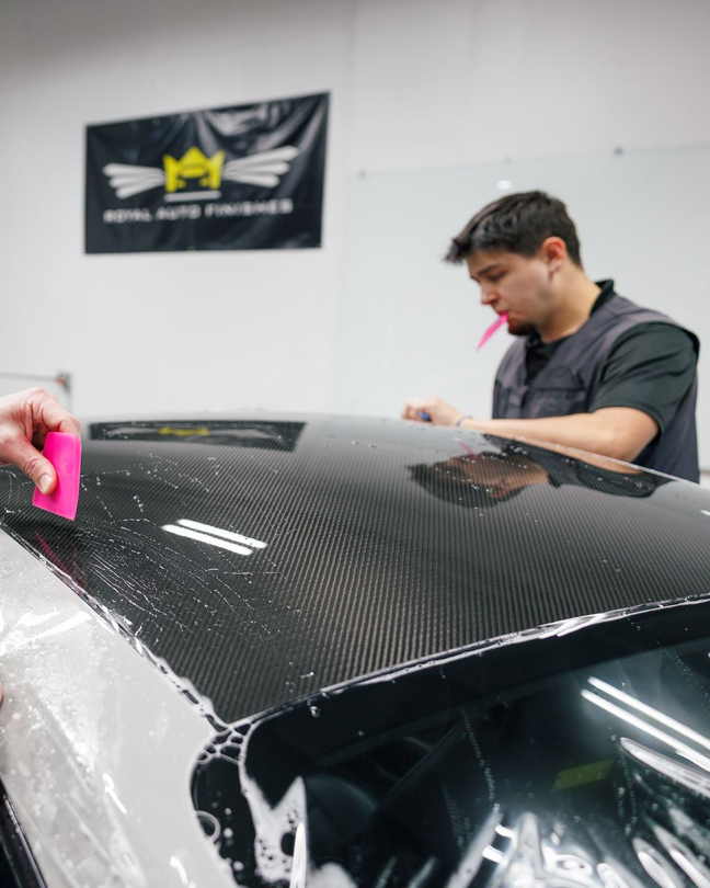 Man applying film to a car roof inside a shop, using squeegee. Royal Auto Finish logo is visible.