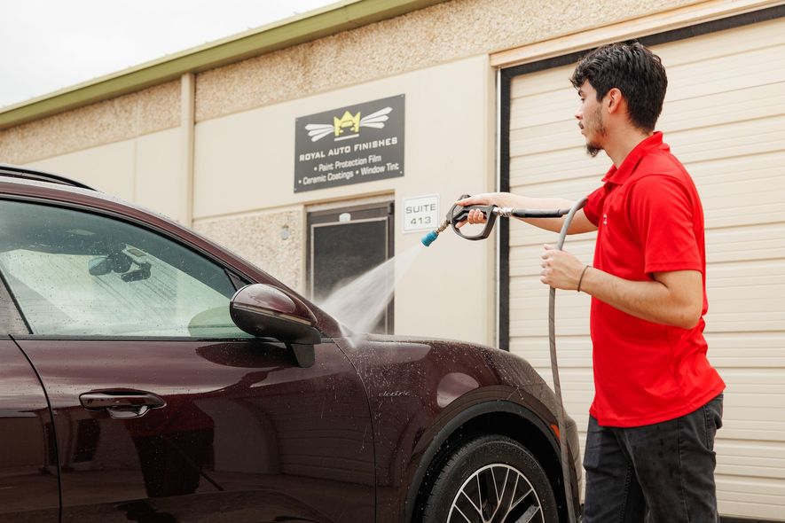 Man in red shirt washing a burgundy car with a power washer outside a building.