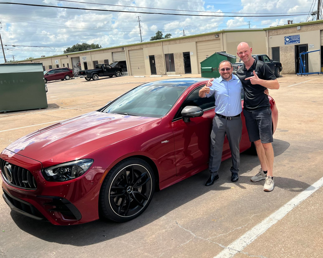 Two men beside a red Mercedes-Benz. One points at car. Building and blue sky in background.