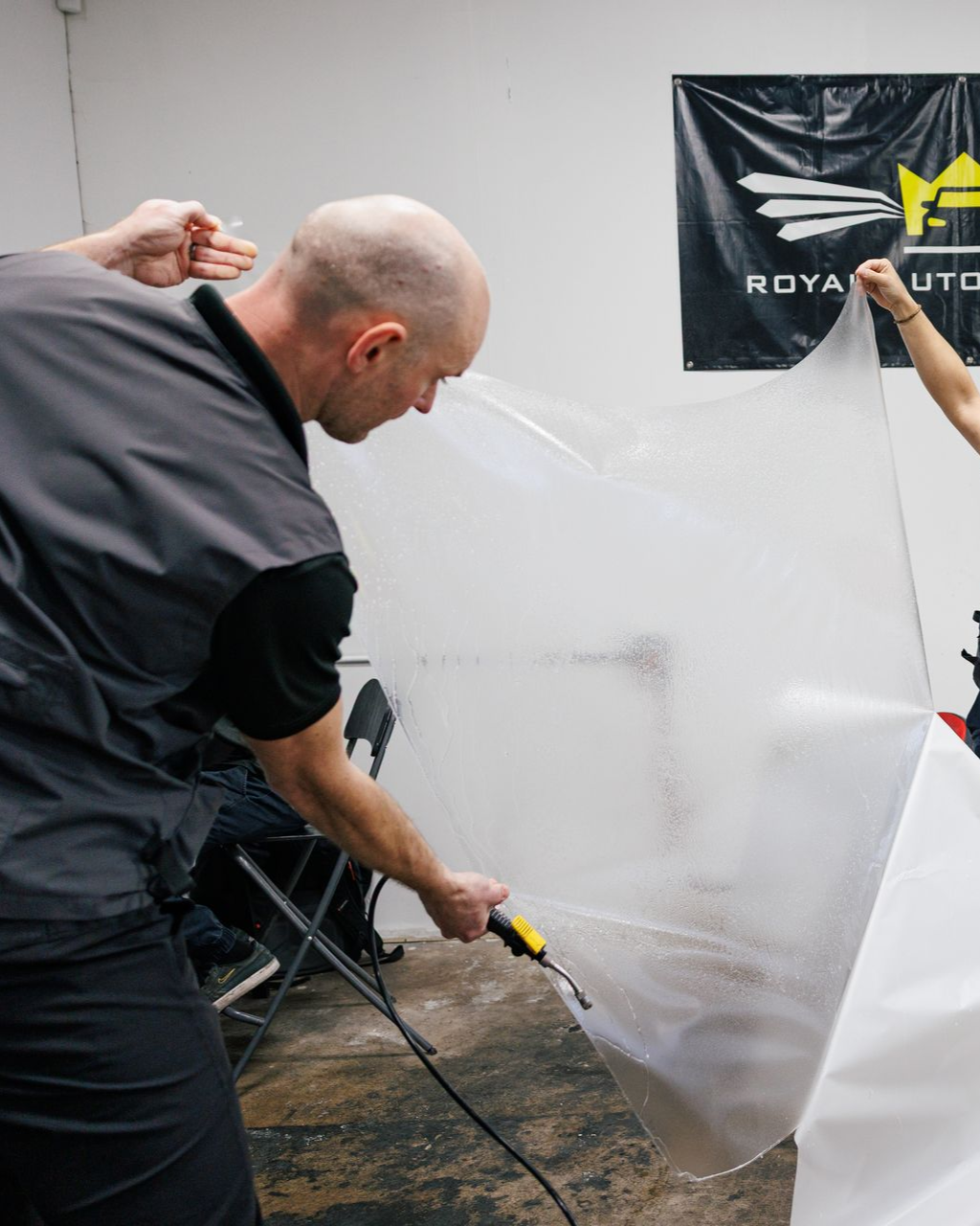 Man applying film to a car, holding a heat gun. Another person holds the other side. Indoor setting.