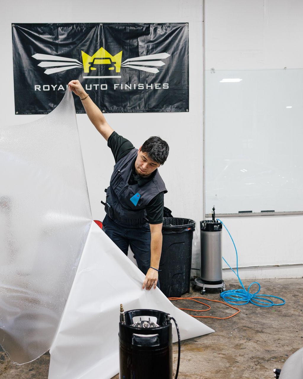 Man preparing to apply a protective film in an auto shop. Holds up large, clear sheet, near a machine.
