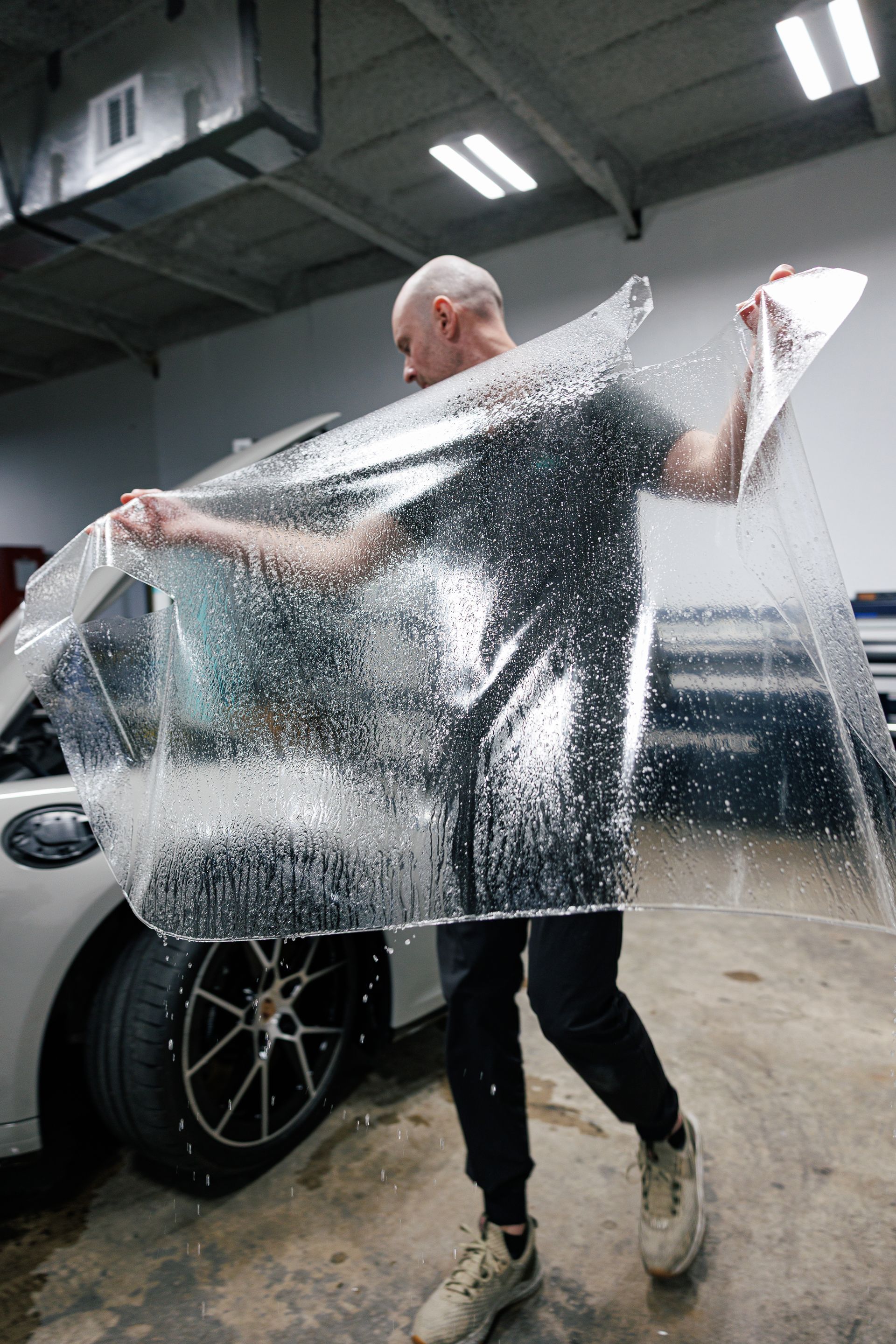 Man holding clear film over a car window, inside a garage.
