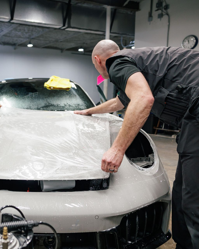 A person applying protective film to a car's front. Gray car, bald man, indoor setting.