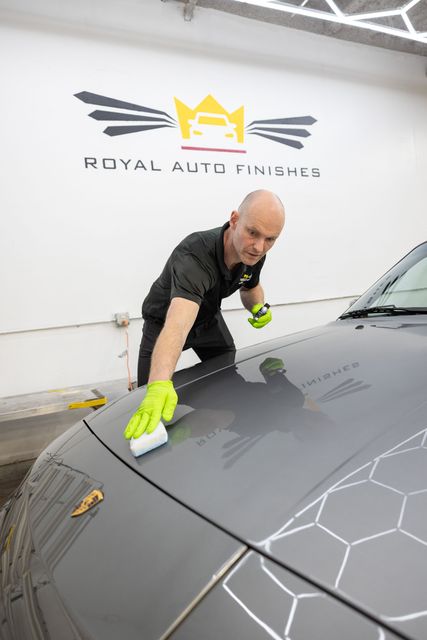 Man applying a sealant to a gray car hood in a detailing shop. He wears gloves and a black shirt.