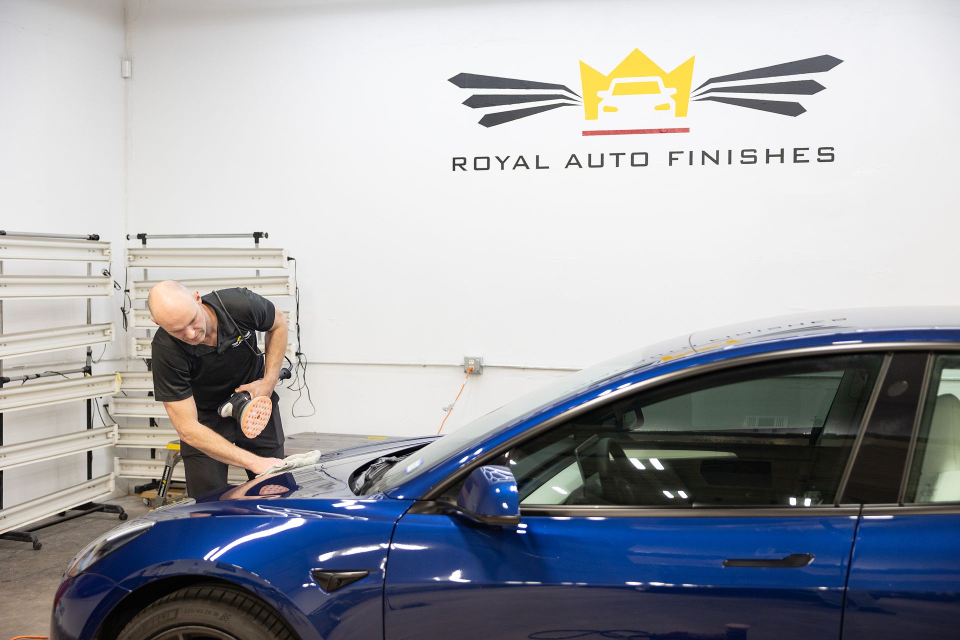 Man working on a blue car in a shop, possibly applying a protective film. "Royal Auto Finishers" logo visible.