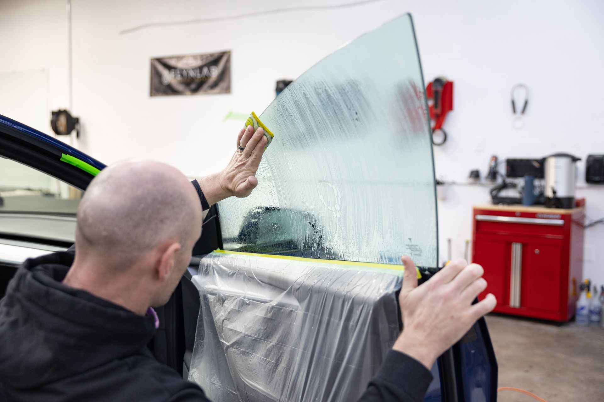 Person applying window tint to a car window in a garage.